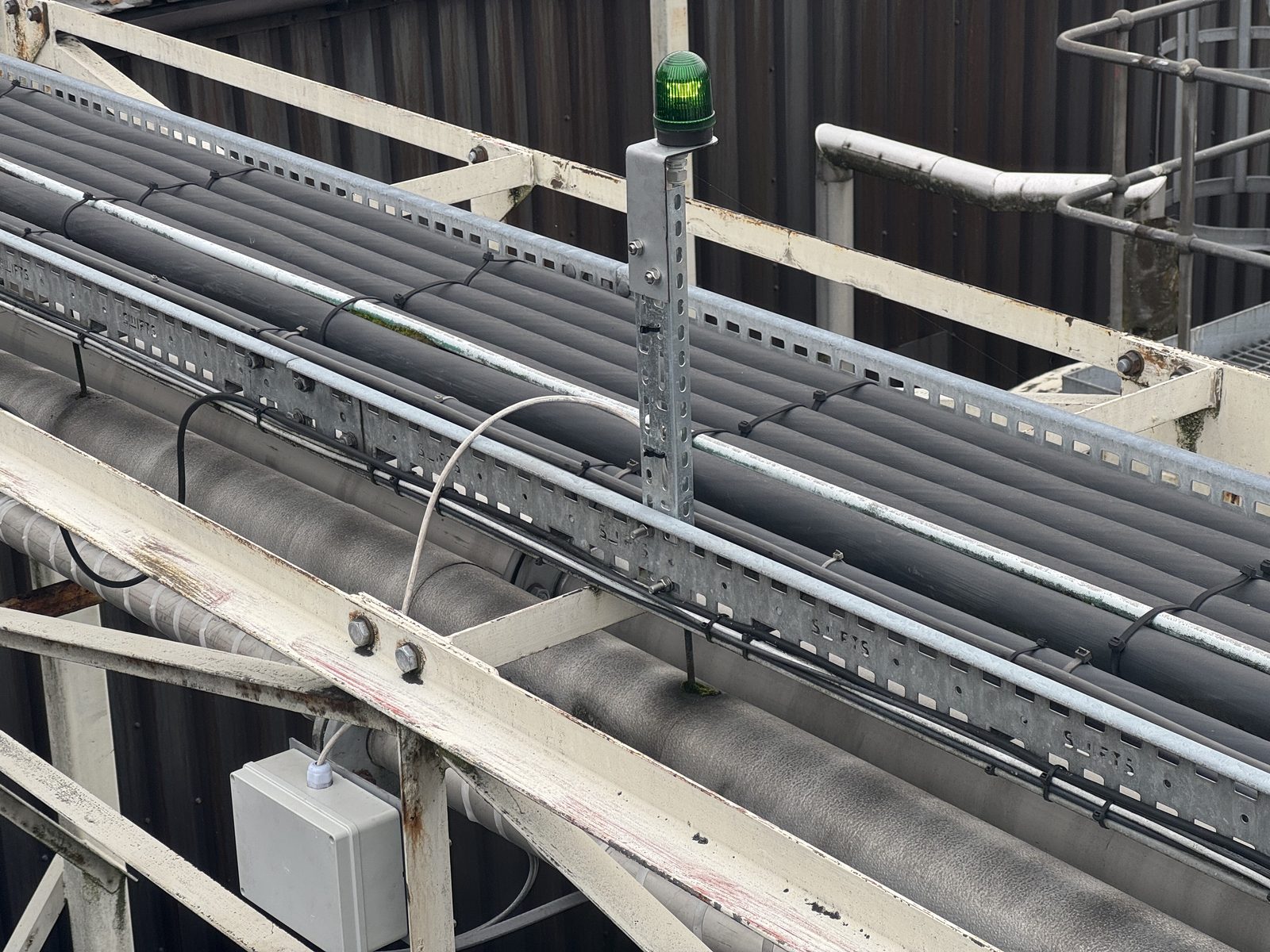Close-up of insulated process pipes on a cable tray with self-regulating heat trace cables zip-tied along them, green beacon light visible