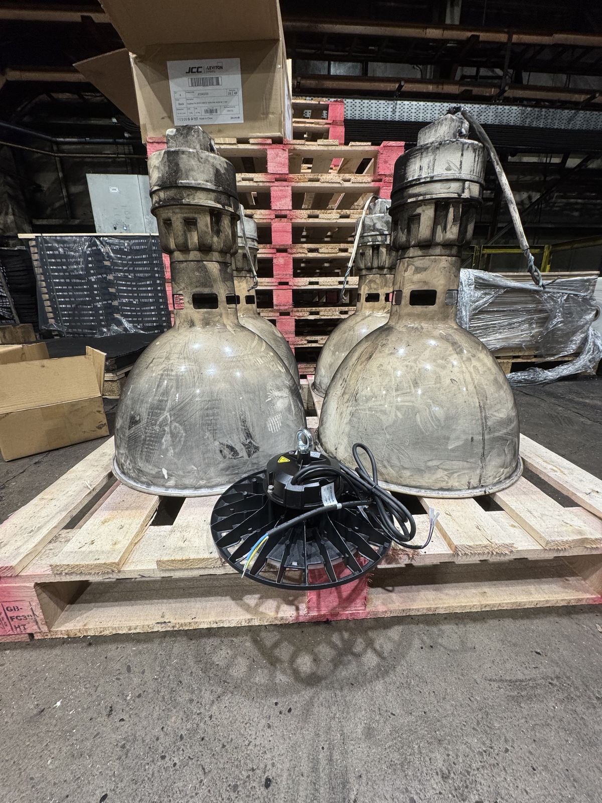 Two old sodium high bay fittings and one new LED high bay on a wooden pallet, with JCC Leviton product box visible in the background
